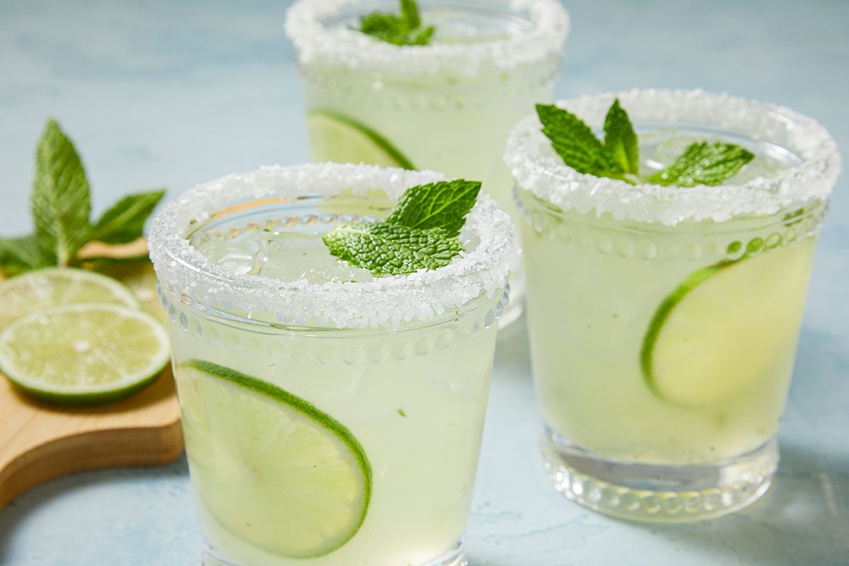 Eye Level horizontal image of three margarita cocktails in glasses with rims coated in sugar. Each glass contains a lime slice, ice, and a mint sprig garnish.