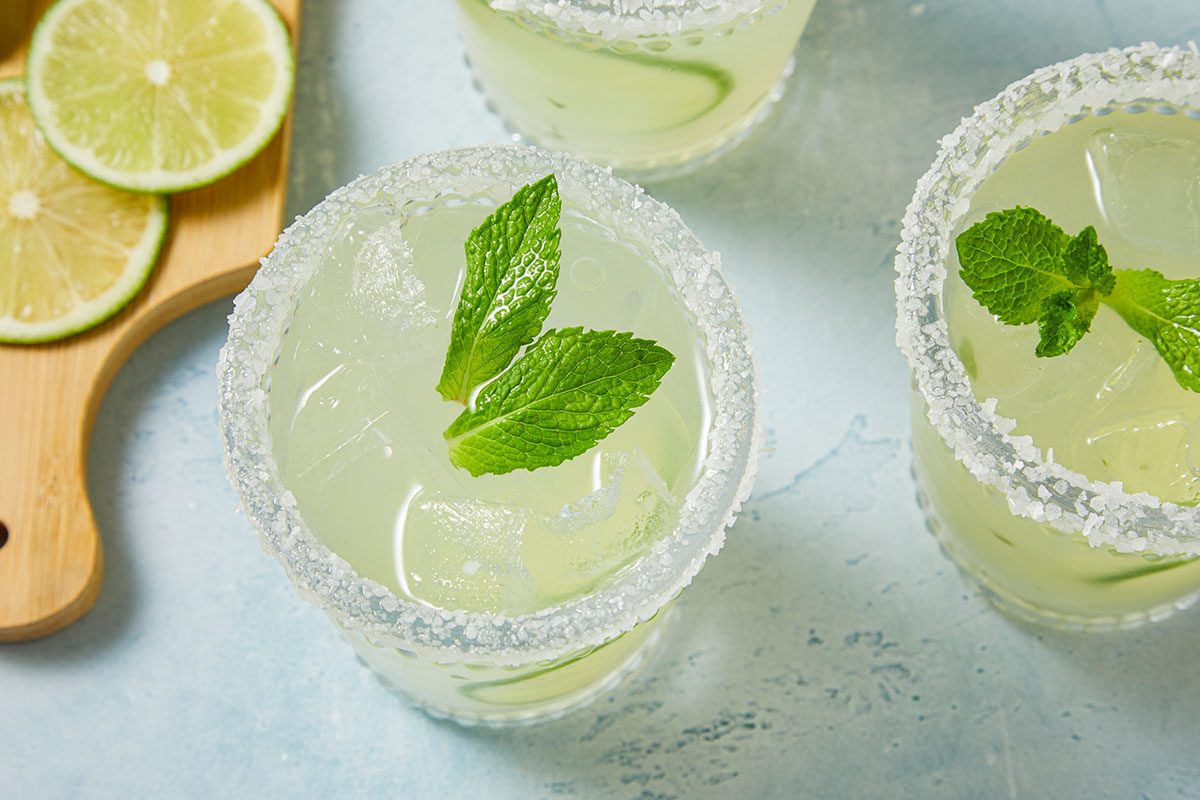 Overhead horizontal image of a set of sugar-rimmed margarita glasses, garnished with mint leaves. The glasses contain ice cubes and lime slices. There are also slices of lime on a wooden board in the corner.