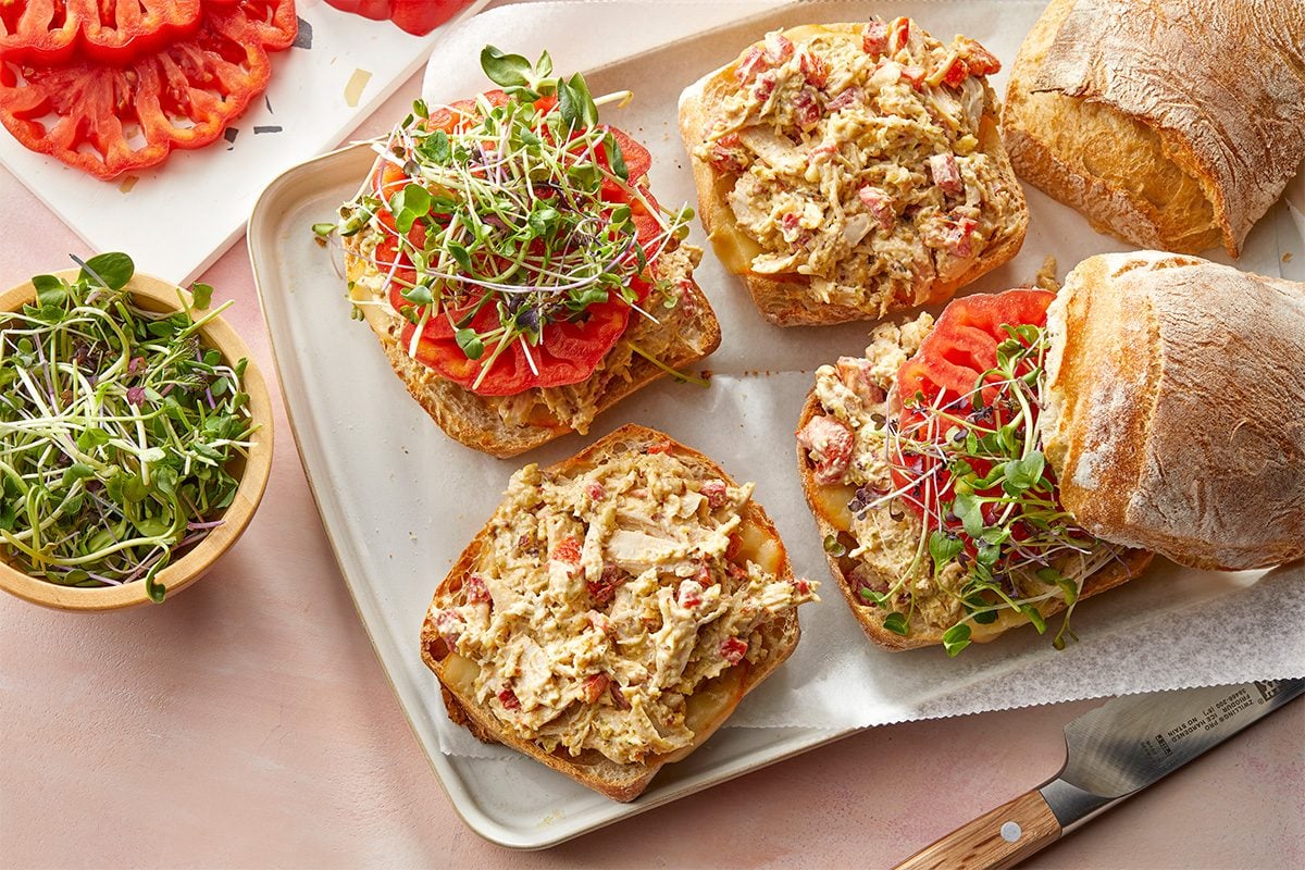 Overhead horizontal image of open faced sandwiches on a metal serving tray. The sandwiches are topped with a chicken salad mixture, sliced tomatoes and sprouts; some have ciabatta bread displayed nearby, and some sliced tomatoes and sprouts are in dishes next to the tray.