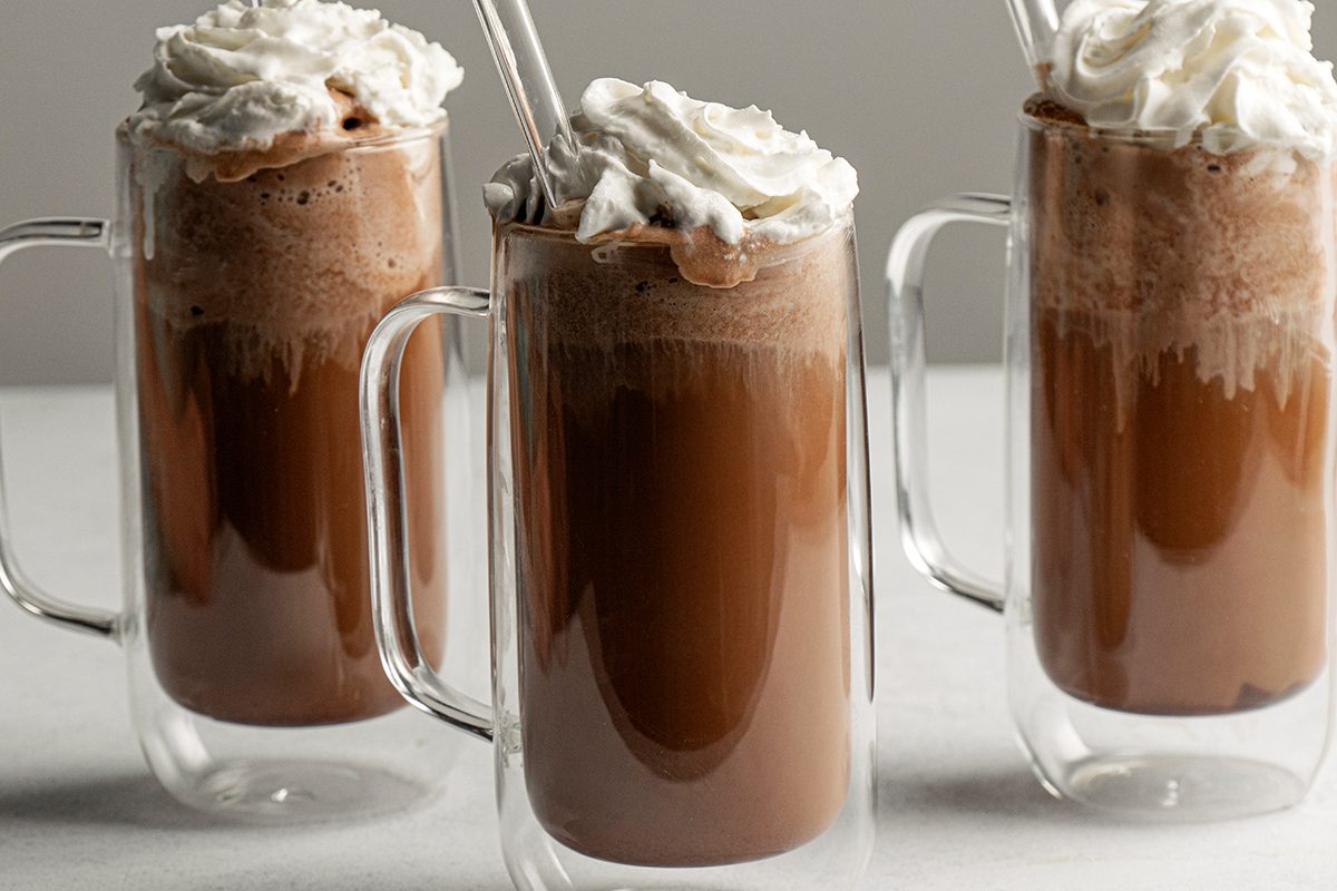 Table view shot of three glass mugs of Old-Fashioned Ice Cream Sodas topped with whipped cream and clear straws, placed on a light surface against a plain background;