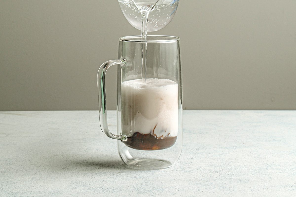 Table view shot of a clear glass mug filled with coffee and milk as water is poured in, creating a swirling effect inside the mug against a plain light background;