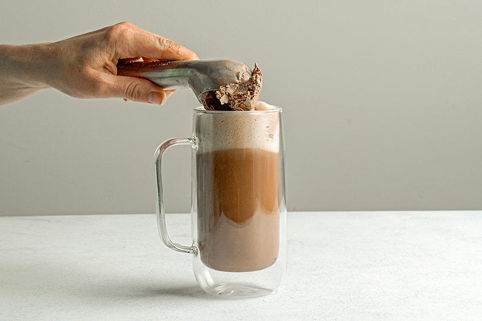 Table view shot of a hand holding an ice cream scoop, placing chocolate ice cream onto a glass mug filled with a frothy brown beverage, against a plain light background;
