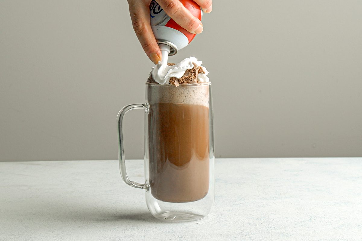 Table view shot of a hand topping a glass mug of hot chocolate with whipped cream from a can, against a light gray background;
