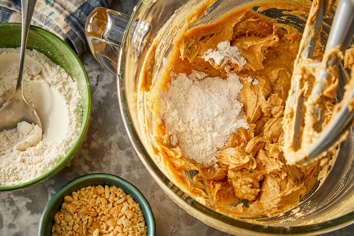 A mixing bowl with peanut butter cookie dough and flour, surrounded by smaller bowls containing flour and chopped nuts, sits on a gray countertop with a spoon and a kitchen towel nearby.
