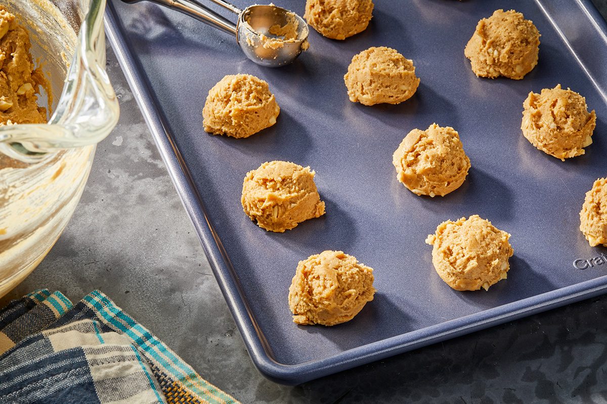 Scoops of cookie dough arranged on a baking sheet, with a mixing bowl, ice cream scoop, and kitchen towels nearby on a countertop.