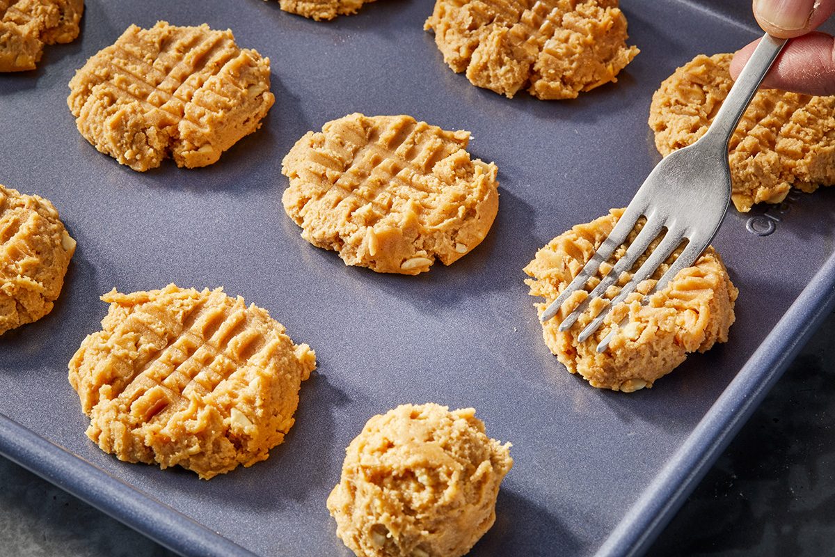 Unbaked peanut butter cookies on a baking sheet, with a hand using a fork to press a crisscross pattern into the dough.