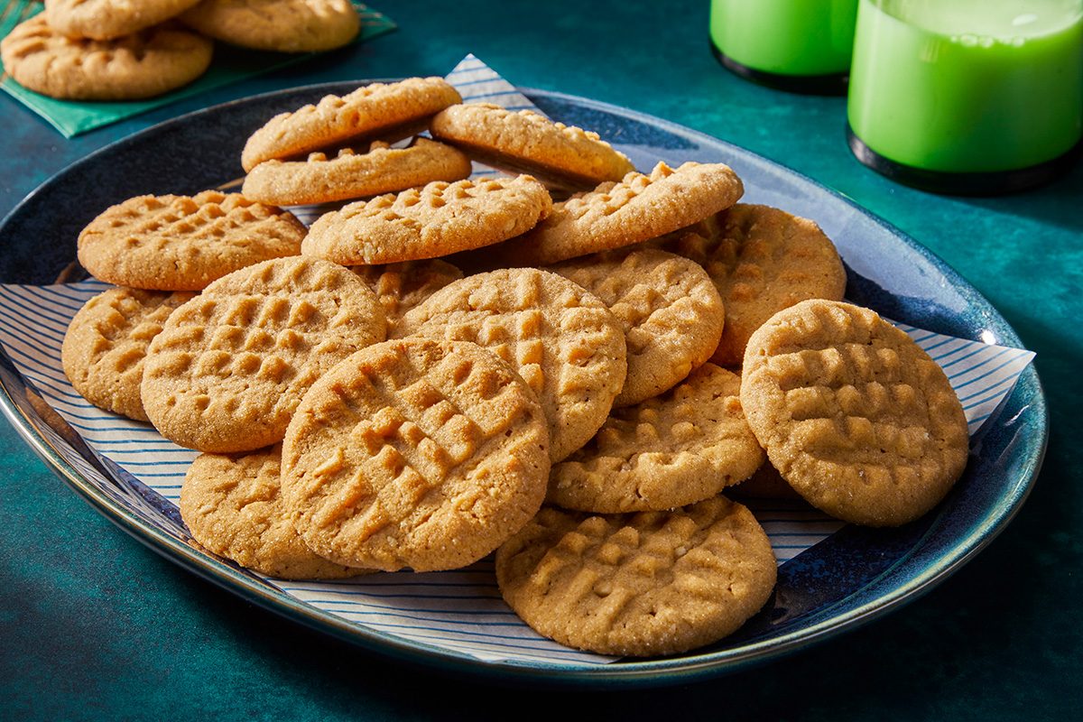 A plate of round peanut butter cookies with crisscross fork marks on top, arranged on a blue plate with a paper liner. Two green glasses with milk are in the background on a green surface.