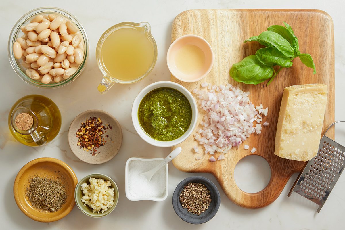 A wooden cutting board with basil leaves, chopped shallots, and a block of Parmesan cheese next to bowls of white beans, broth, pesto, olive oil, spices, chopped garlic, and grated cheese on a white surface.