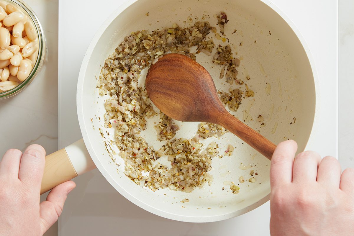 A person stirs chopped onions and herbs in a white skillet with a wooden spoon. A bowl of white beans is nearby on the counter.