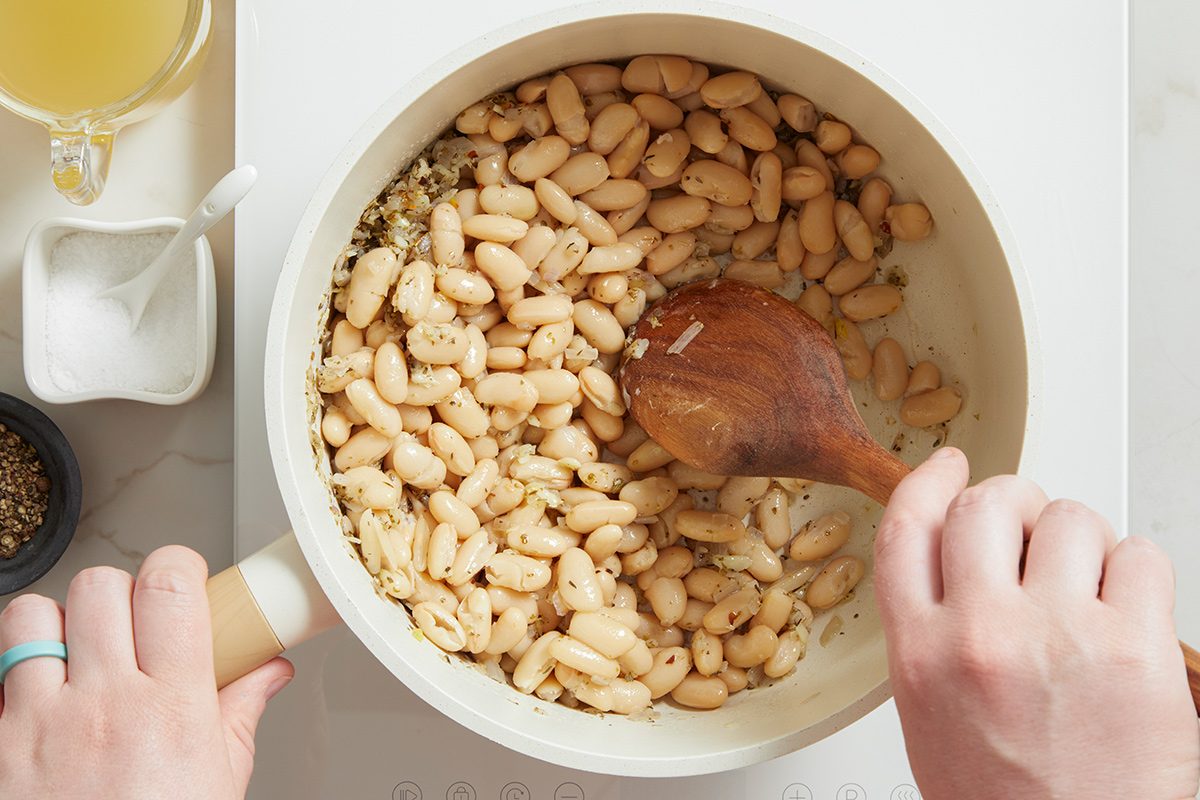 A person stirs white beans with a wooden spoon in a white pot on a stove. Small bowls of salt, pepper, and broth are placed nearby.