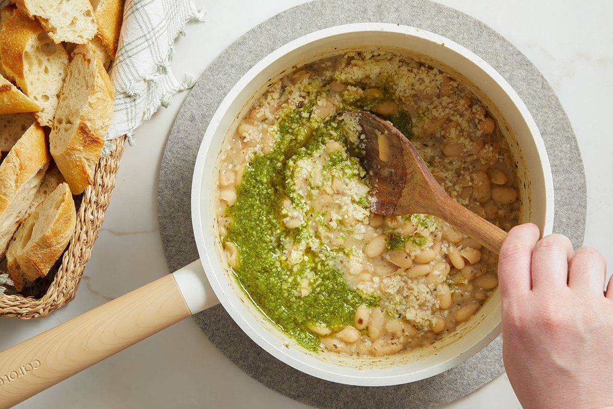 A hand stirs white beans topped with green pesto in a saucepan, with a basket of sliced bread on the side.