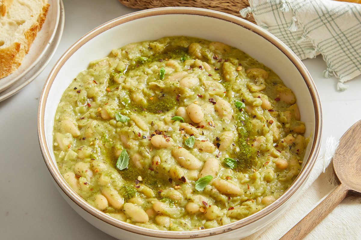 Bowl of thick white bean stew with herbs and olive oil, garnished with fresh green herbs, placed on a light tabletop with a wooden spoon and a checkered napkin nearby.