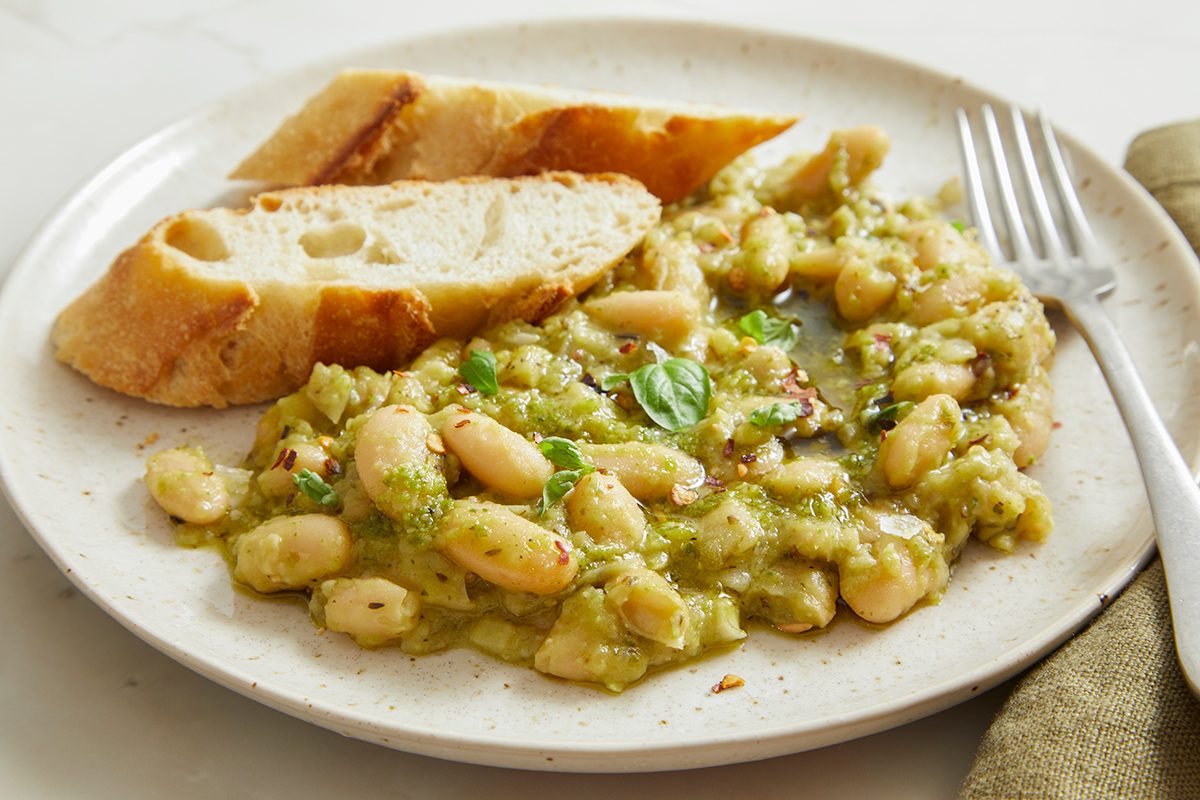A plate of white beans in a green herb sauce garnished with parsley, served with two slices of crusty bread and a fork on the side.