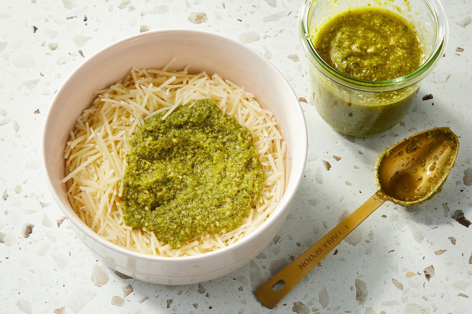 A large batch of shredded Parmesan topped with some pesto sauce in a medium bowl. A spoon and more pesto placed on side.