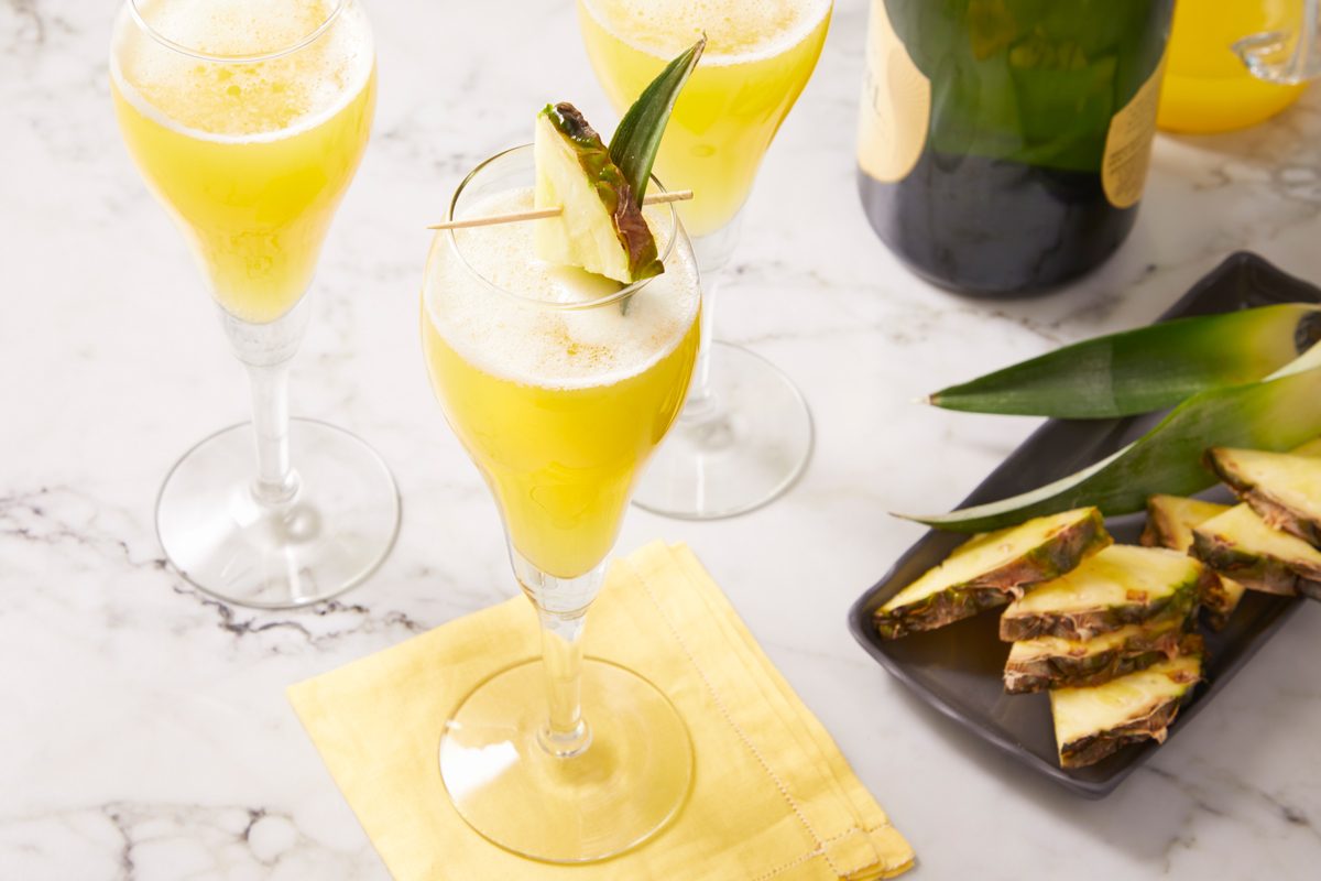 Eye Level horizontal image of three flute glasses filled with a yellow cocktail garnished with pineapple on a marble surface. A bottle of champagne and a black plate with pineapple chunks and leaves are in the background.