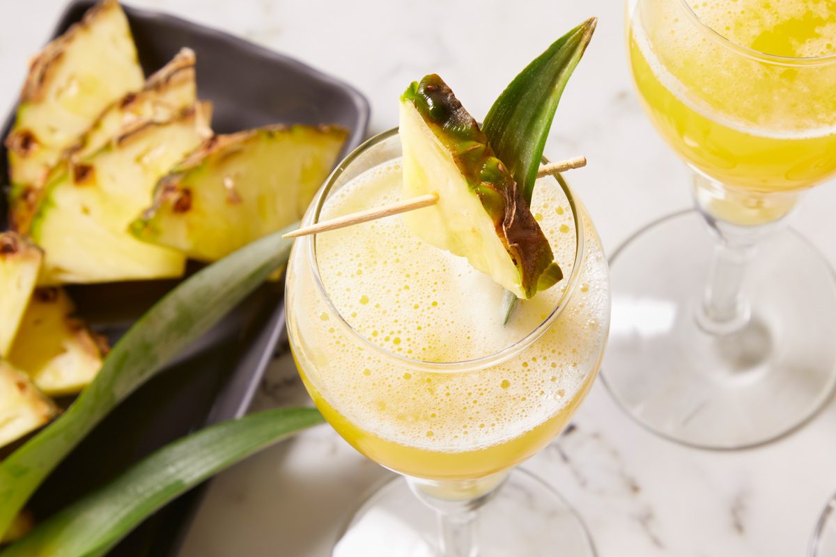 Overhead horizontal image of a tropical pineapple cocktail. Pineapple wedges are served on a black tray and one cocktail is garnished with a pineapple wedge and leaf on a toothpick.