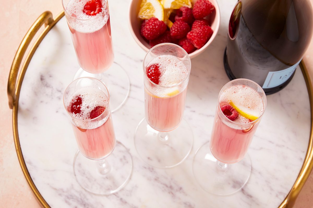 Four champagne flutes with pink sparkling drinks, garnished with raspberries and lemon slices, on a marble tray with a gold rim. A bowl of raspberries and a bottle are also on the tray.