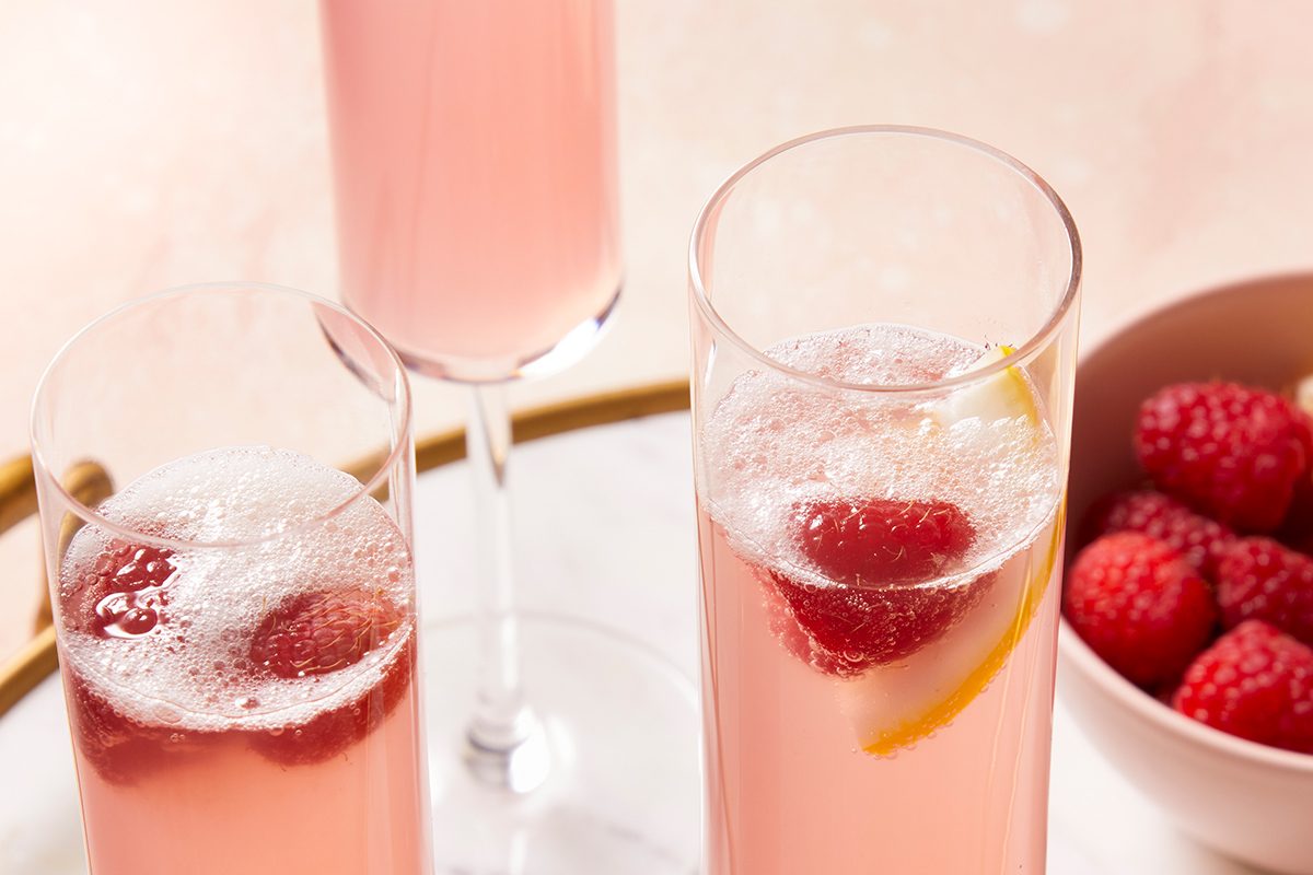 Three glasses of pink sparkling drinks with raspberries and a lemon slice, next to a bowl filled with fresh raspberries, all placed on a light surface.