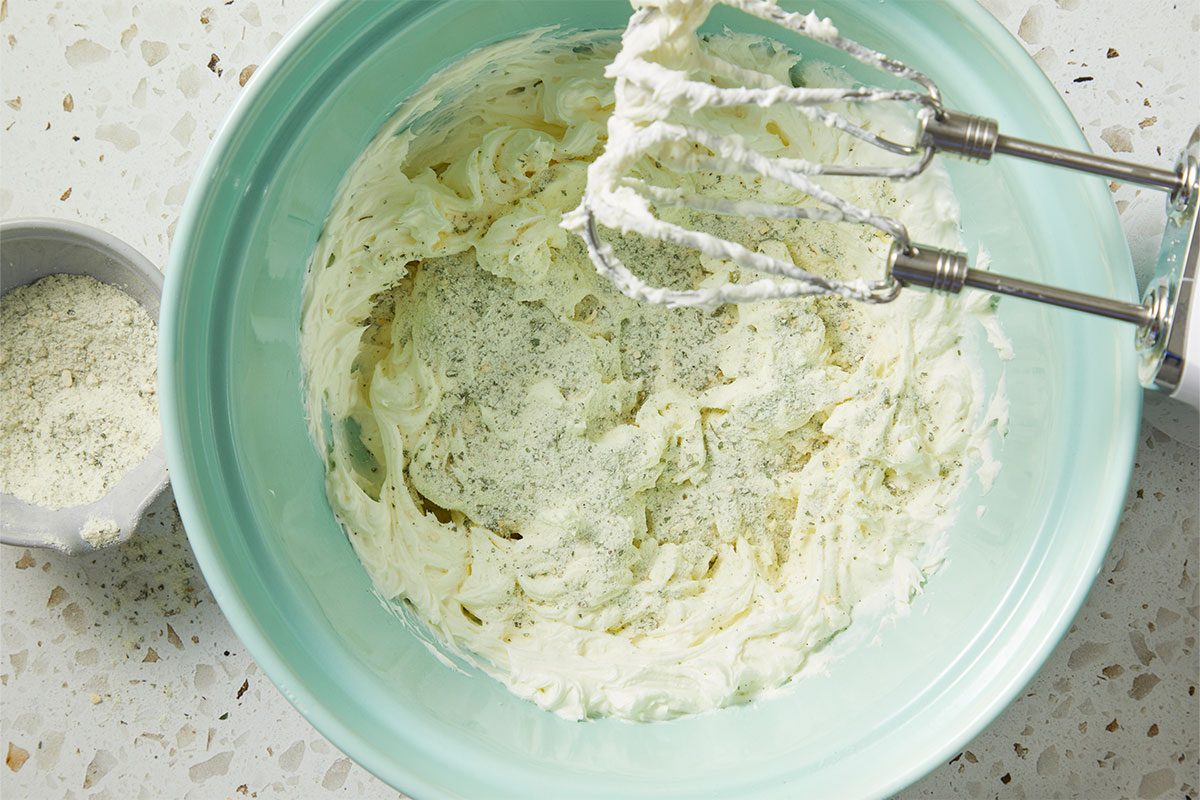 Overhead horizontal image of cream cheese in a light blue mixing bowl with seasoning and an electric mixer. A bowl of shredded cheese is visible to the side.