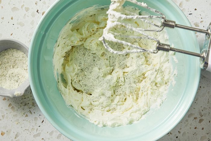 Overhead horizontal image of cream cheese in a light blue mixing bowl with seasoning and an electric mixer. A bowl of shredded cheese is visible to the side.
