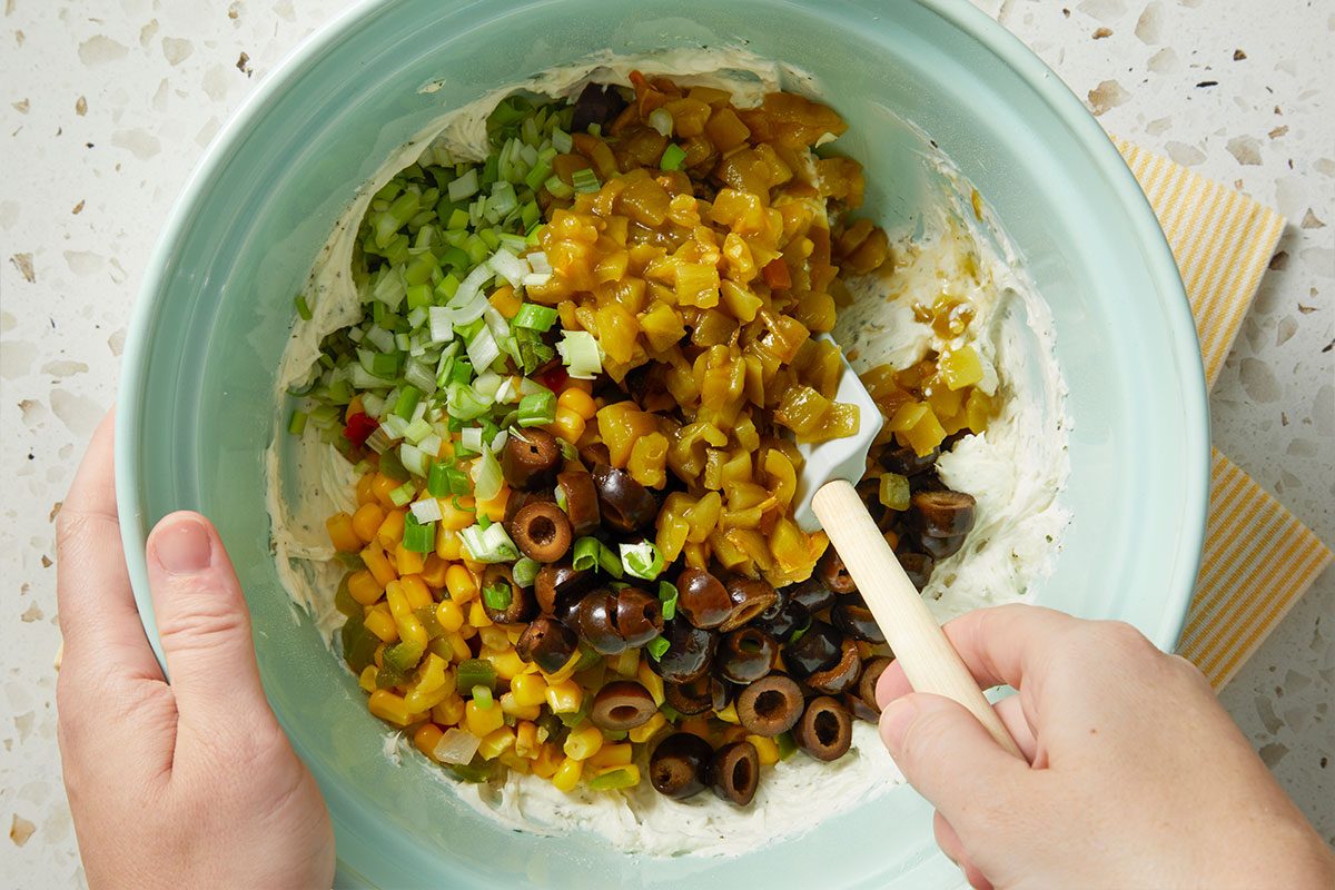 Overhead horizontal image of ingredients being added to a bowl of cream cheese. Corn, olives, green onions, and peppers are being added to the bowl with a spatula.