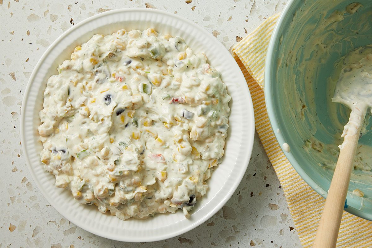 Overhead horizontal image of a bowl of corn dip with black beans and peppers, next to a blue bowl with a spoon. The bowl of dip is placed on a white plate on a speckled countertop.