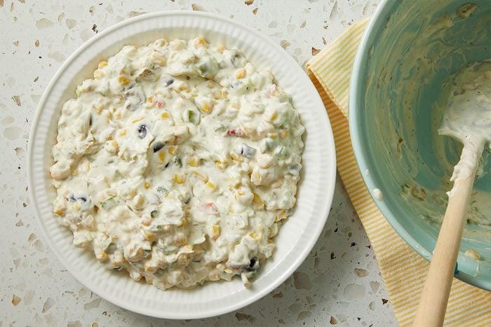 Overhead horizontal image of a bowl of corn dip with black beans and peppers, next to a blue bowl with a spoon. The bowl of dip is placed on a white plate on a speckled countertop.