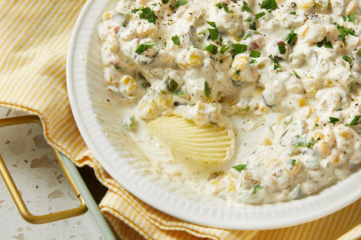 Overhead horizontal image of a bowl of corn dip, garnished with parsley and pepper. A ruffled potato chip is dipped into the cream based corn dip, featuring corn, green peppers, and onions.