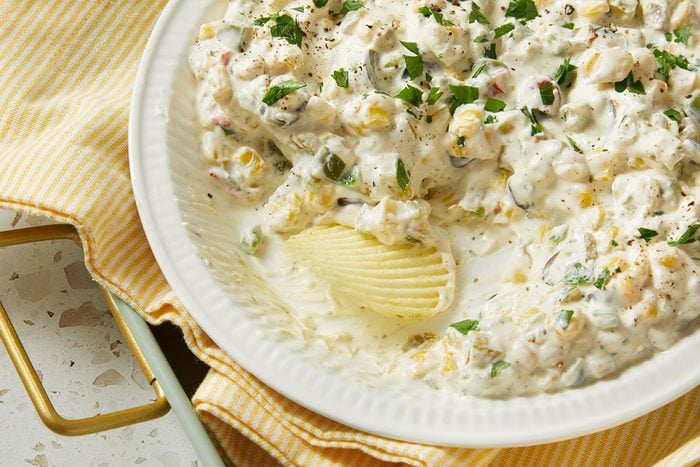 Overhead horizontal image of a bowl of corn dip, garnished with parsley and pepper. A ruffled potato chip is dipped into the cream based corn dip, featuring corn, green peppers, and onions.
