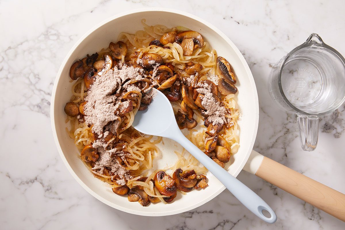 Overhead horizontal image of onions and mushrooms sautéing in a skillet with seasoning.