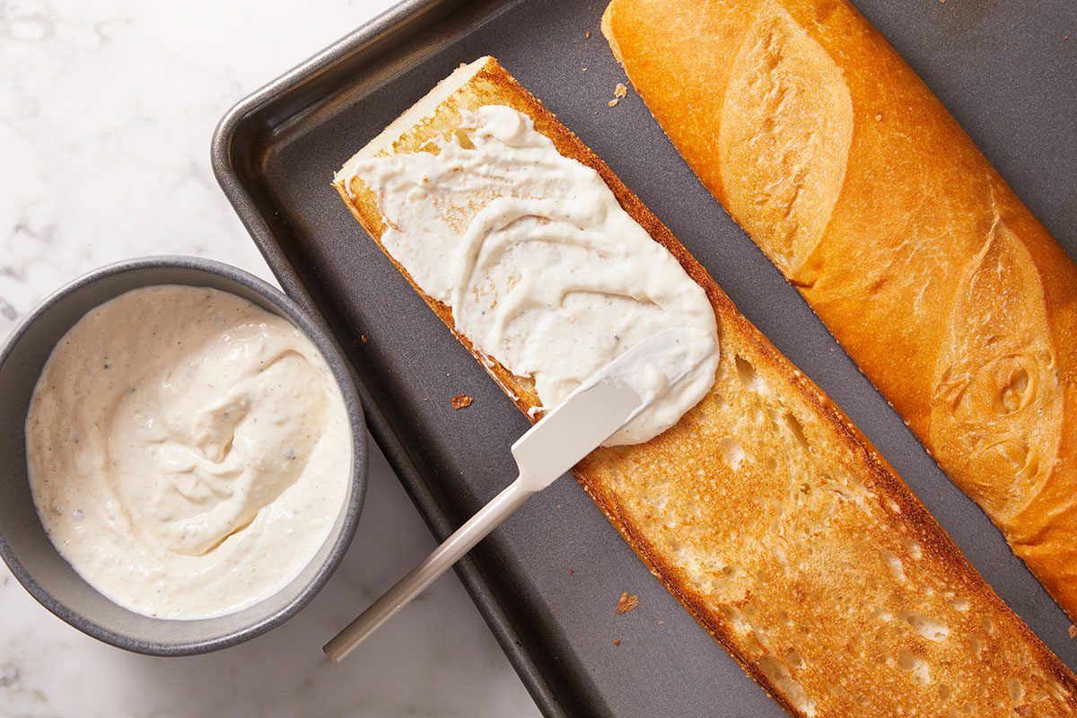 Overhead horizontal image of mayonnaise and horseradish spread being applied to toasted French bread.