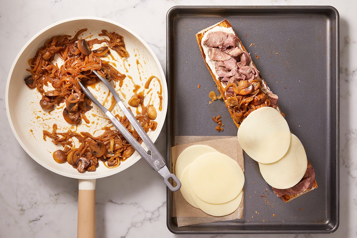 Overhead horizontal image of sandwich assembly with sliced prime rib, sautéed onions, mushrooms, and cheese.