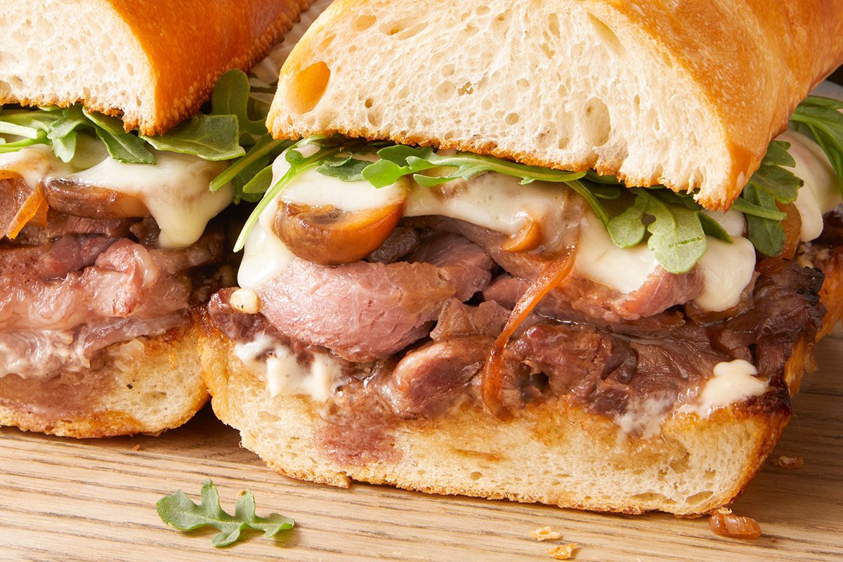 3/4 angle horizontal close-up of prime rib sandwich showing layers of beef, cheese, and arugula.