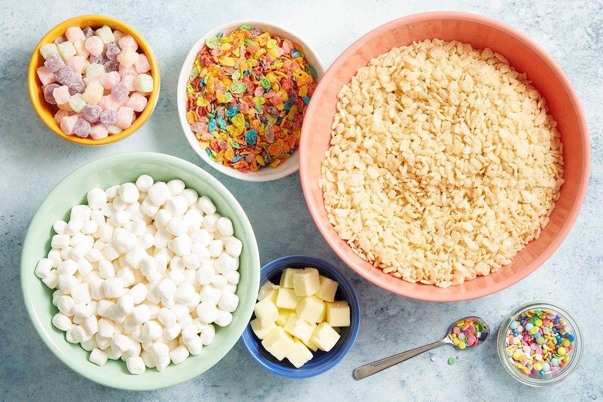 Five bowls filled with colorful gumdrops, Fruity Pebbles cereal, Rice Krispies cereal, mini marshmallows, and butter cubes, with a small spoon holding rainbow sprinkles on a light surface.