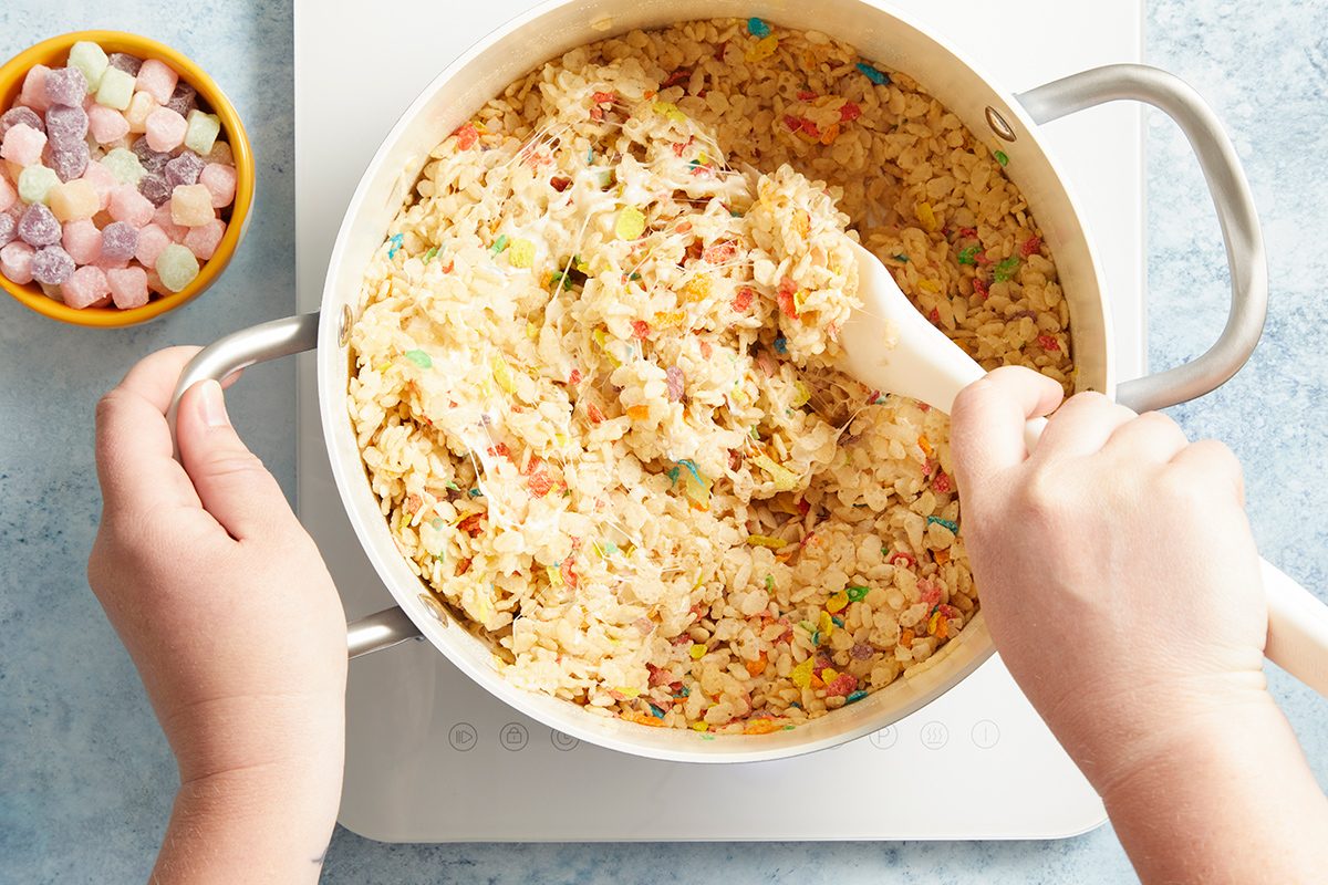 A person stirs colorful rice cereal treat mixture in a white pot with a white spoon. A small bowl of pastel marshmallows sits nearby on a light blue surface.