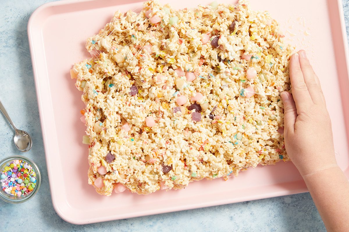 A hand presses a mixture of rice cereal and colorful marshmallows onto a pink tray, with a spoon and a small bowl of rainbow sprinkles nearby on a light surface.