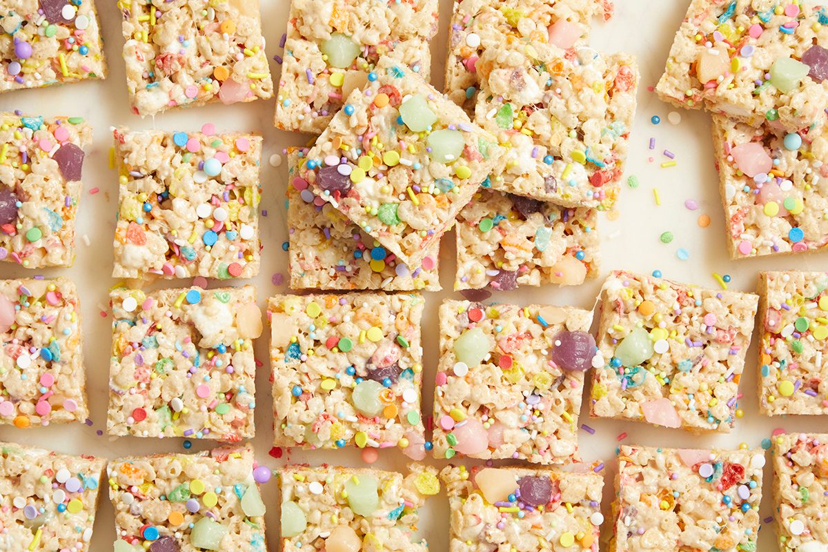 Squares of rice crispy treats topped with colorful sprinkles and candy decorations are arranged closely together on a light surface.