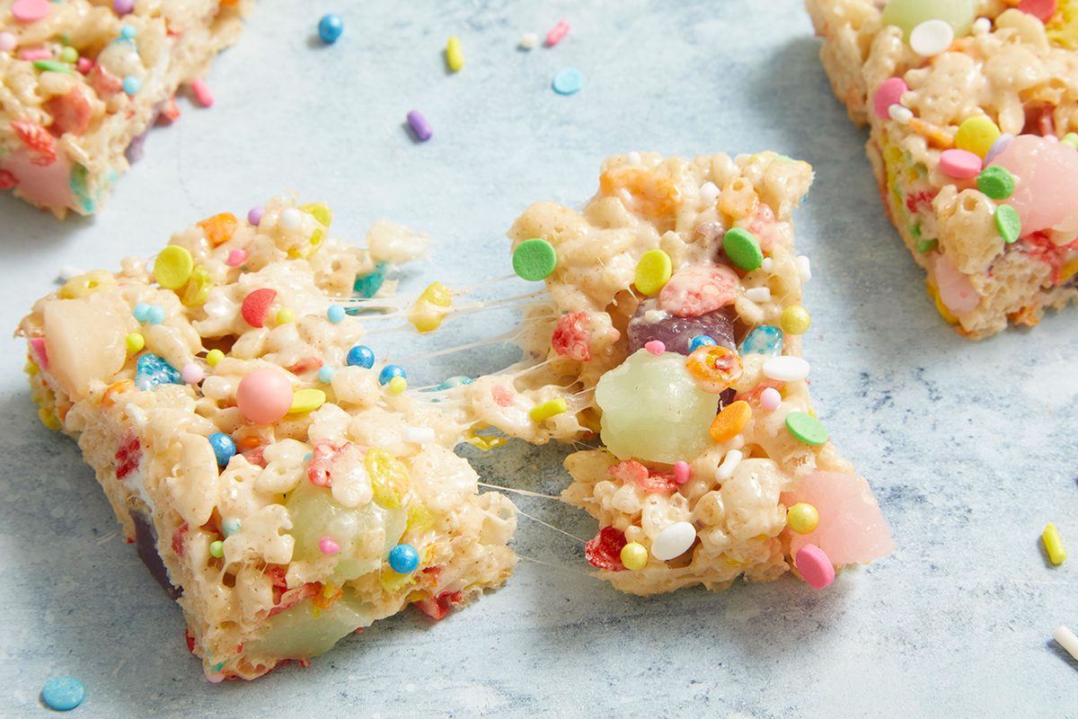 Colorful crispy rice treats with marshmallows and rainbow sprinkles are shown on a light blue surface. Two pieces are pulled apart, with gooey marshmallow stretching between them.