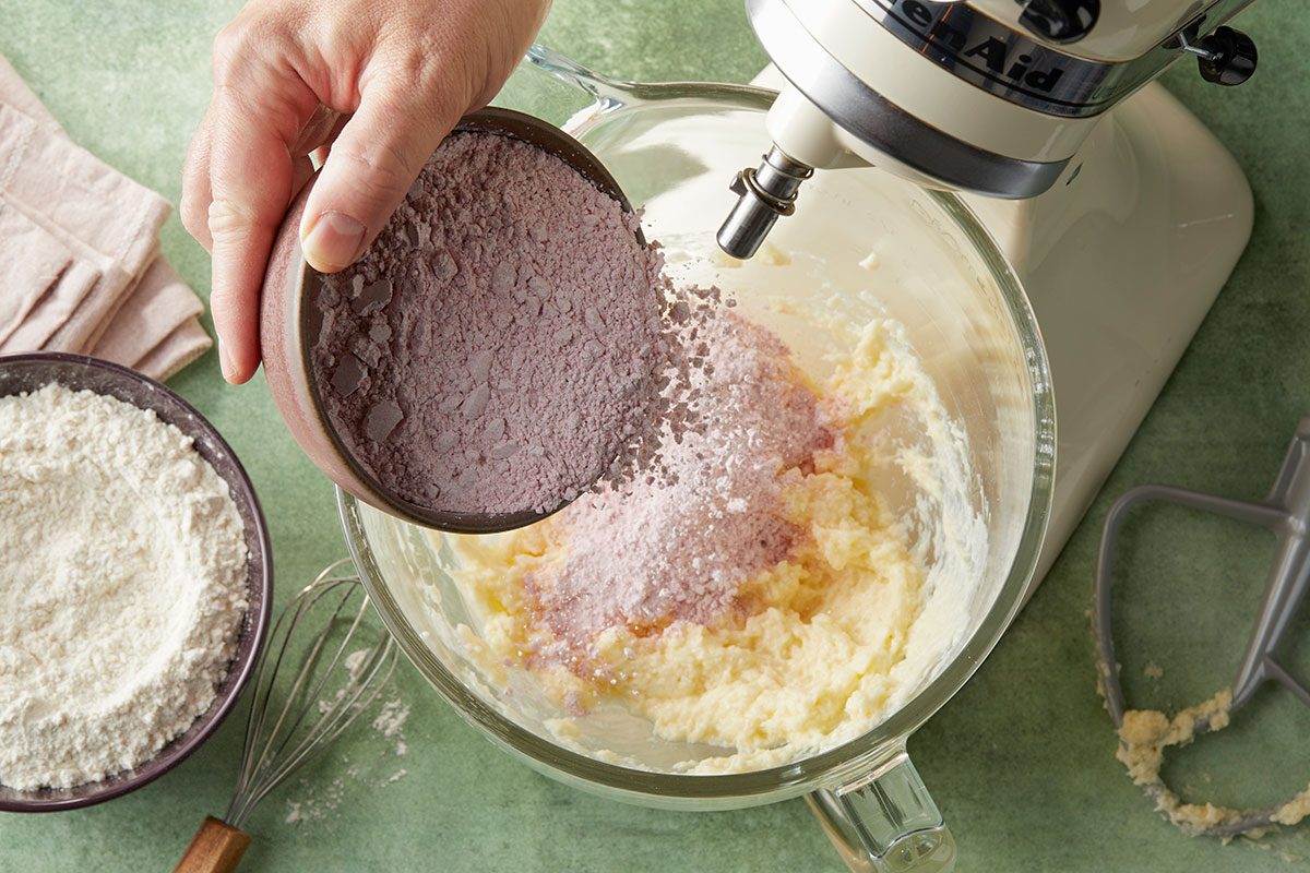 Overhead horizontal image of adding dry ingredients from a bowl into a bowl of batter that is mixed with a KitchenAid stand mixer. There is a bowl of flour next to the bowl of batter, along with a whisk. | [Original] Adding confectioners’ sugar with raspberry powder to creamed butter mixture, bowl with flour mixture on the side ready to add next