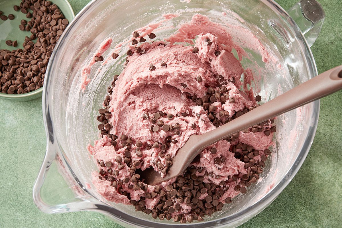 Overhead horizontal image of cookie dough in a glass mixing bowl with chocolate chips being folded in using a silicone spatula. Additional chocolate chips sit in a small green bowl to the left. | [Original] Stirring chocolate chips into batter