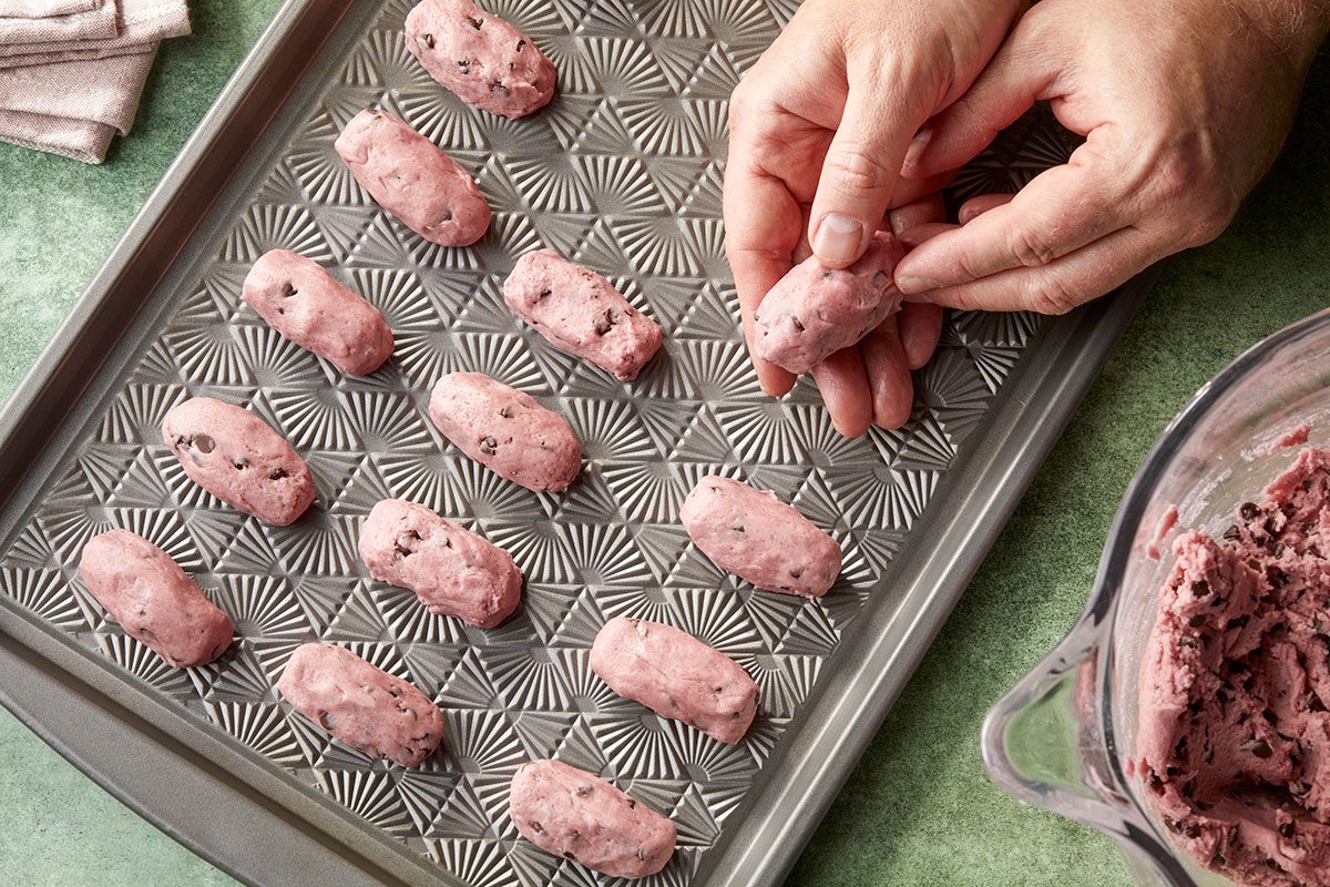 Overhead horizontal image of cookie dough being shaped. Hands are forming dough into an oval shape. Some dough has already been placed on the baking sheet. | [Original] Forming dough into logs and placing on baking sheet