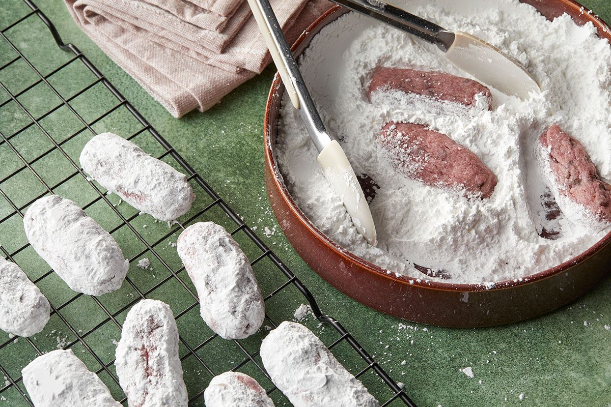 Overhead horizontal image of small logs being coated in powdered sugar inside of a bowl with the help of tongs. A cooling rack with logs already coated with sugar is in view. | [Original] Rolling baked cookies in confectioners’ sugar
