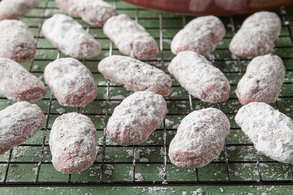 Overhead horizontal image of cookies dusted with powdered sugar, cooling on a black wire rack. The cookies have a slightly oblong shape.