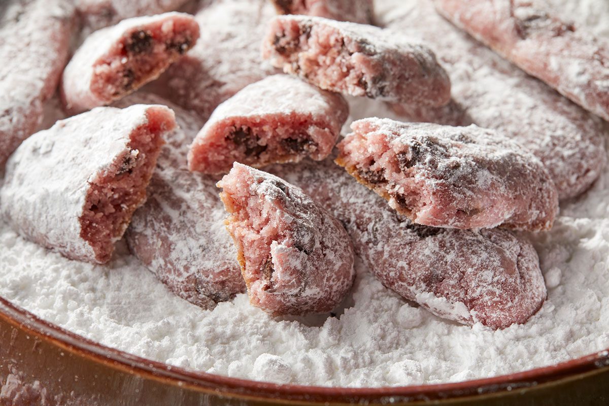 Eye Level horizontal image of chocolate cherry cookies coated with powdered sugar. The cookies have a light pink hue, and one is broken open to reveal the interior.