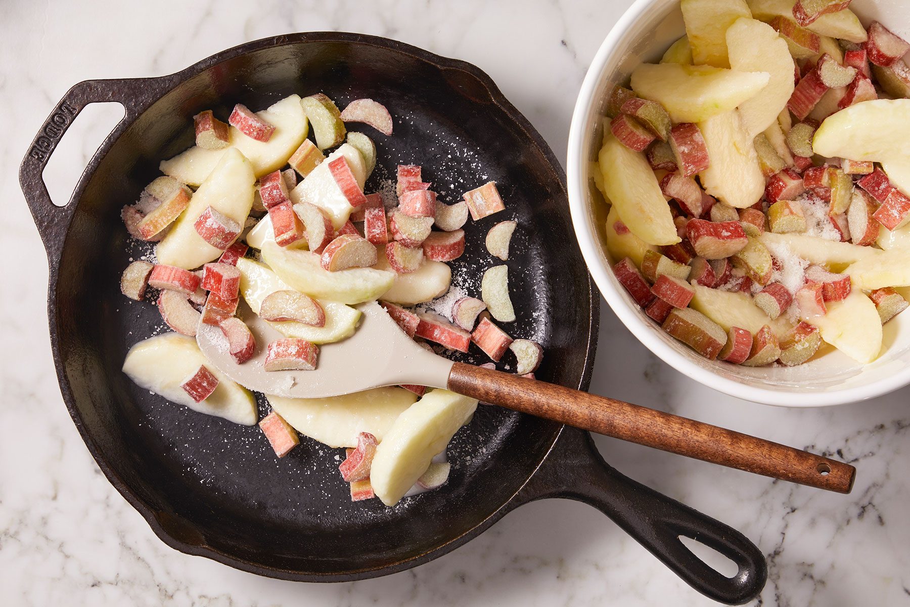 Chopped rhubarb and apples with cornstarch and sugar in a large iron skillet with a bowl of rhubarb and apples in a small white bowl.