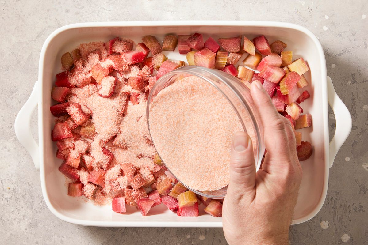 A horizontal overhead image shows chopped rhubarb in a baking dish as sugar and gelatin mixture is being poured over the fruit.