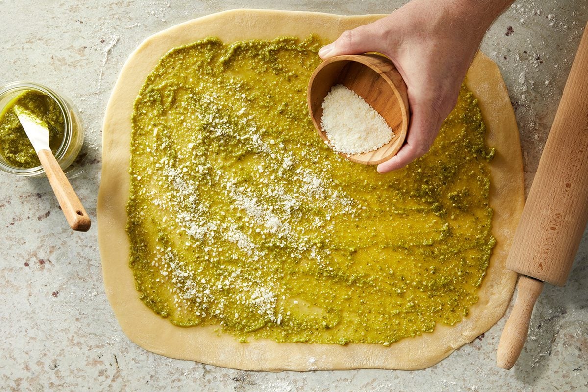 Overhead horizontal image of dough slathered with pesto sauce and Parmesan cheese being added from a small wooden bowl. A jar of pesto is to the left, and a rolling pin to the right. | [Original] RMS 282446 Savory Babka with Pesto; How to; Step 2 Sprinkling grated Parmesan over rolled dough that has been spread with pesto