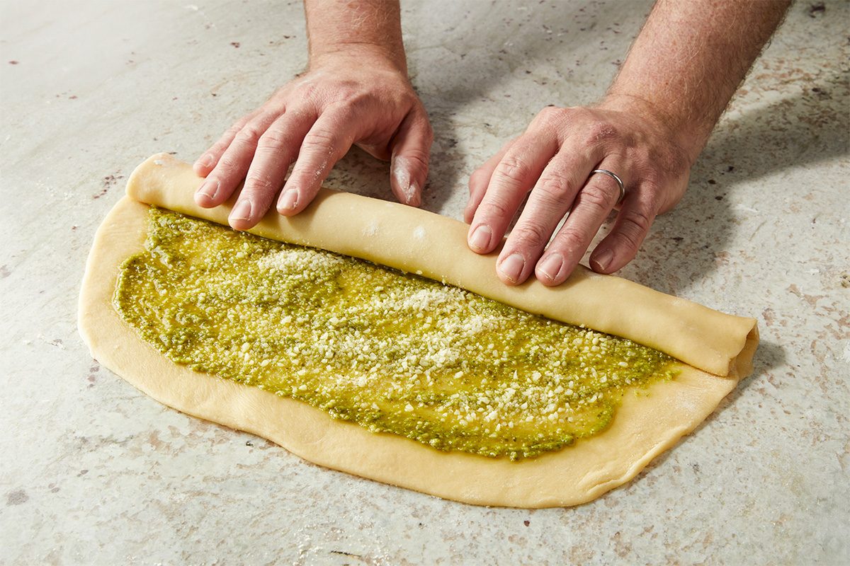 Overhead horizontal image of two hands rolling up dough filled with pesto and topped with parmesan cheese. The dough is being rolled on a neutral colored countertop. | [Original] RMS 282446 Savory Babka with Pesto; How to; Step 3 Rolling dough; jellyroll style