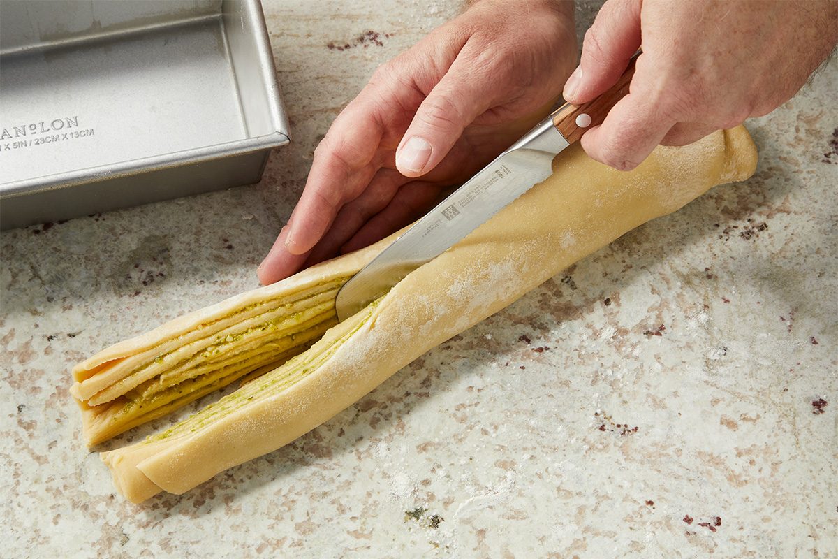 Eye Level horizontal image of a person using a knife to cut a log of layered dough filled with a green pesto-like substance. A metal baking pan is visible in the background. | [Original] RMS 282446 Savory Babka with Pesto; How to; Step 4 Cutting rolled dough in half lengthwise