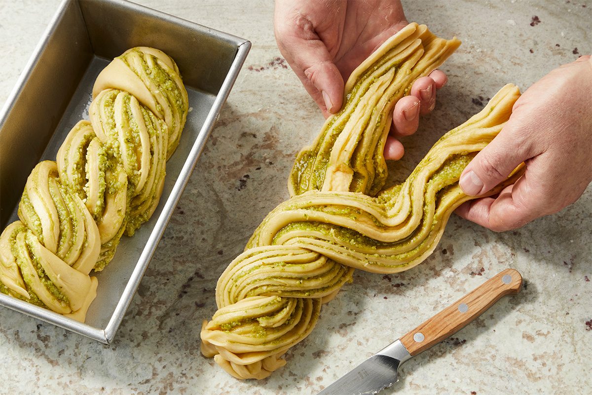 Eye Level horizontal image of the baker holding the dough which has been swirled with pesto. The dough is being placed into a metal loaf pan and there is a paring knife lying on the counter next to the dough. | [Original] RMS 282446 Savory Babka with Pesto; How to; Step 5 Twisting dough; first loaf already twisted and placed in 9x5 pan